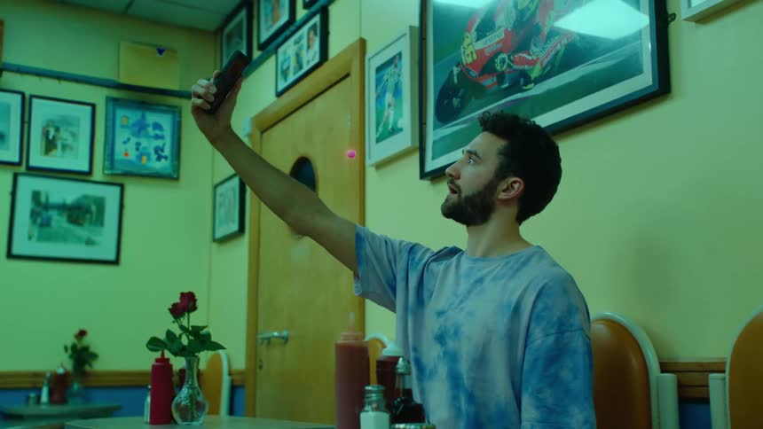 A bearded man wearing a blue tshirt holds up a smartphone to take a selfie and an orange orb floats around