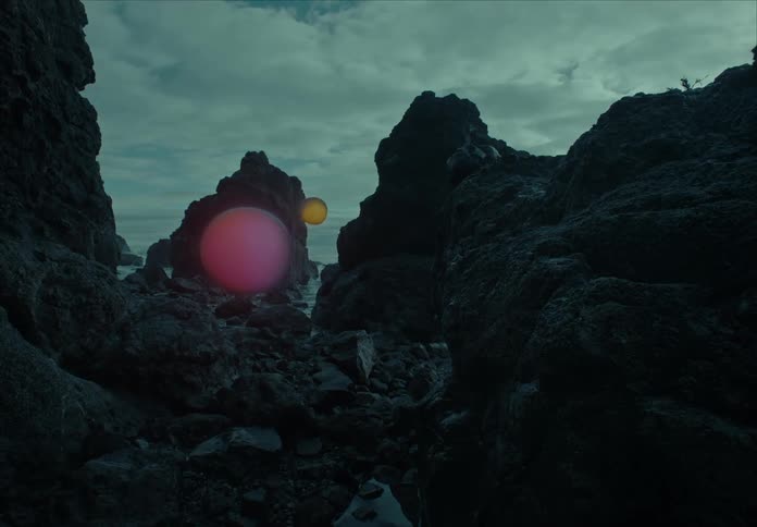 Two round shapes one purple and one orange float between a coastal rock formation while changing colors