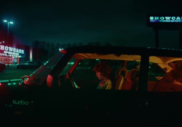A person with curly hair sits in a car looking at a smartphone in a parking lot at night