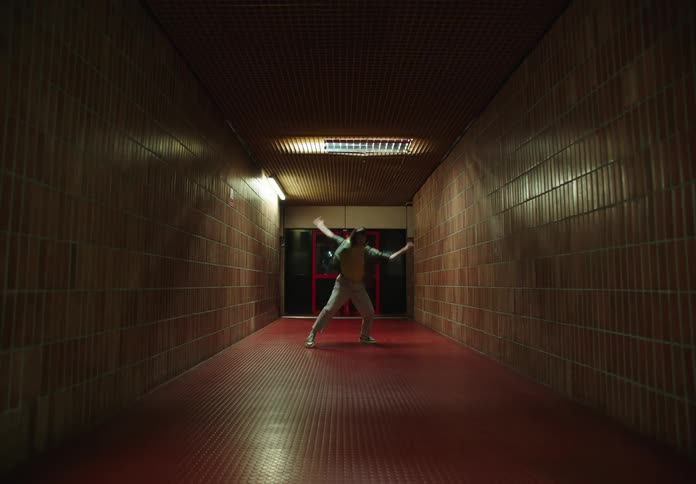 Woman wearing a yellow top and a blue shirt and beige pants dances in a dimly lit corridor