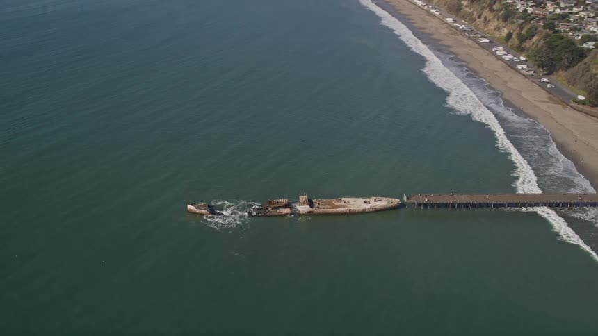 View from above of a pier on a beach reaching a sunken ship stranded in the middle of the sea