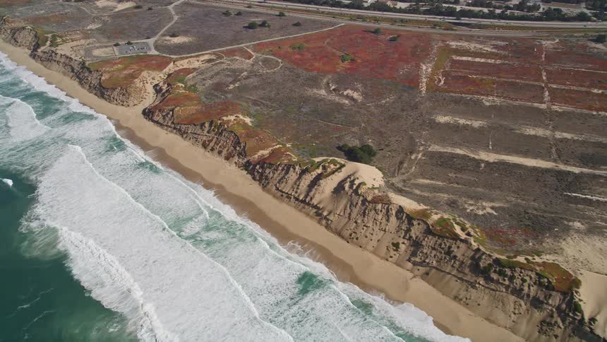 The aerial view shows a beach with dramatic cliffs waves crashing on the shore and a winding path leading up the hillside