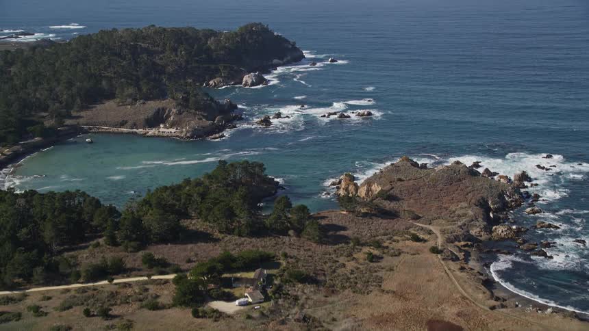 An aerial view of vehicles driving on a coastal road near a rocky coastline with crashing waves