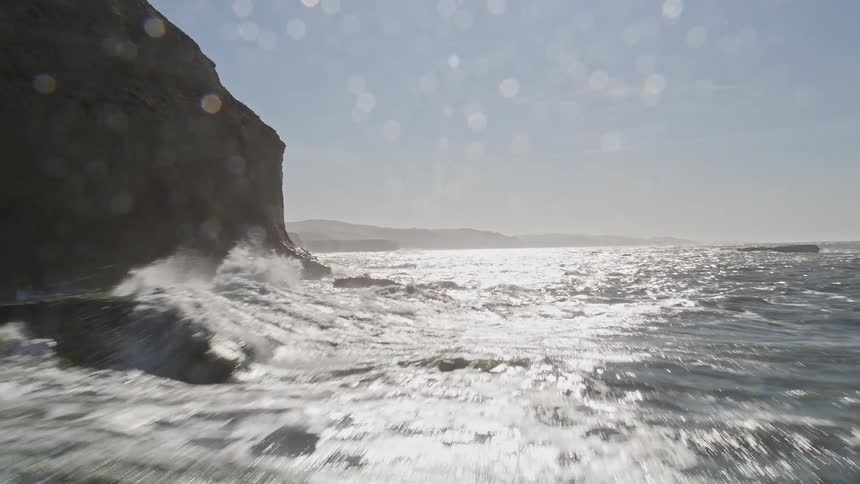 Waves roll onto coast crashing against mountains side cliffs on a sunny day