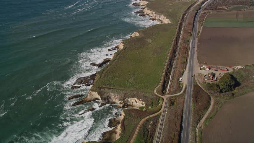 Aerial view of a coastline with waves crashing on the beach next to a cliff and a road parallel to the train tracks