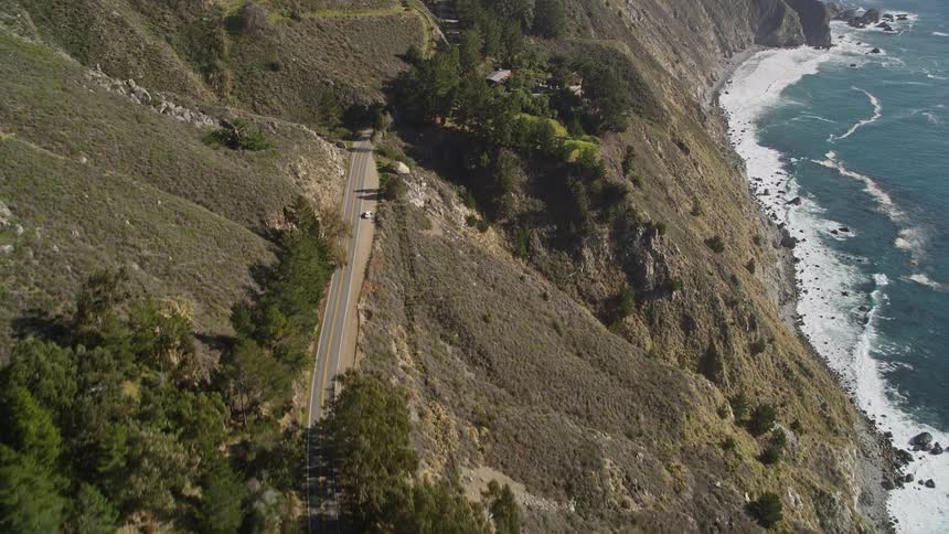 Aerial view of a winding road that cuts along a steep cliff with vegetation and next to the ocean with waves crashing on the shore
