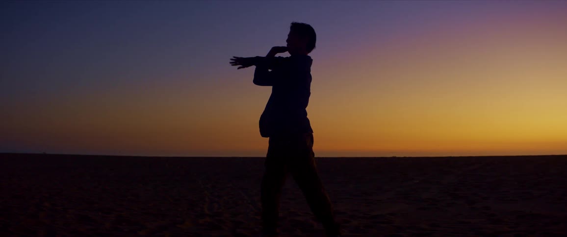 Silhouette of a person dancing moving their arms on the beach sand against a blue and reddish sky