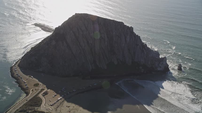 Vehicle drives along coastal highway from the parking lot near mountain casting a shadow onto the beach