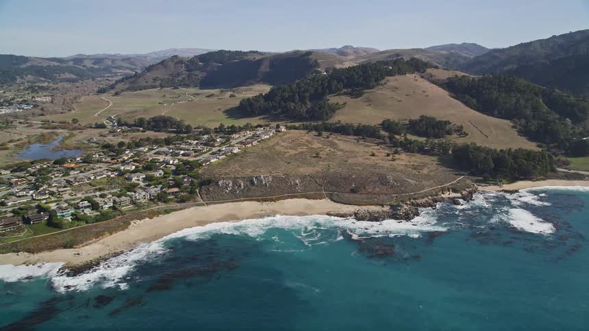 An aerial view shows a beach with turquoise water meeting lush green mountains in the distance
