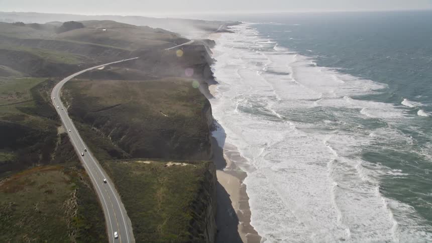 Aerial view of cars driving along a scenic coastal road with dramatic cliffs and crashing waves