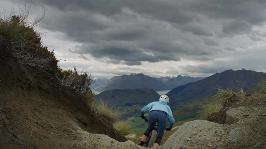 Two mountain bikers are captured midair while performing a jump on a rugged trail set against a backdrop of dramatic cloudy skies and mountainous terrain