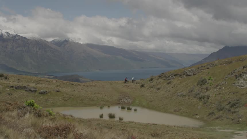 Two bikers biking near a muddy pond and mountains in the center of a hilly area