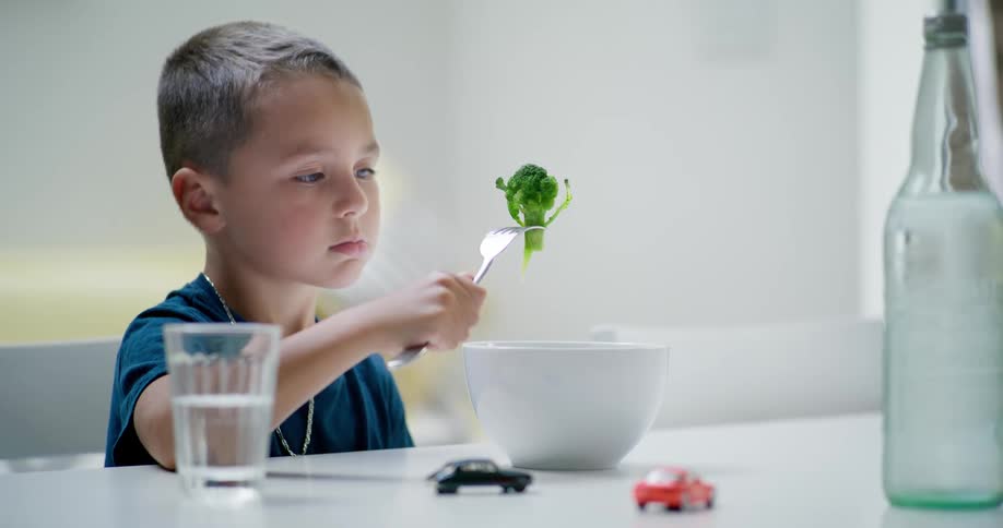 A child sitting has a broccoli on his fork and is trying to eat it but cant