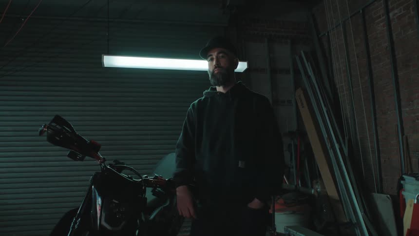 Man with a cap and beard indoors leaning on a motorcycle