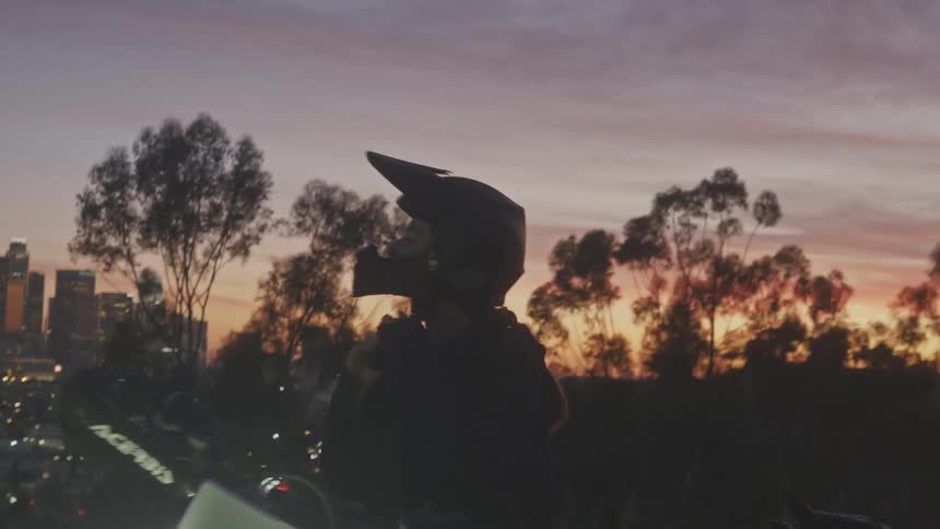 Woman removing her helmet while standing next to her motorcycle on a hill with a panoramic view of a metropolis at sunset