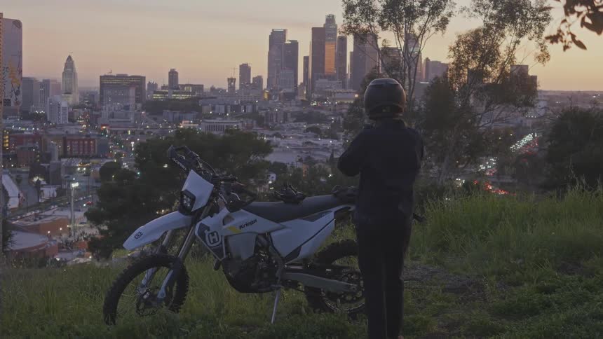 Woman on a hill taking off her helmet and standing next to a motorcycle overlooking a metropolis with large buildings in the late afternoon