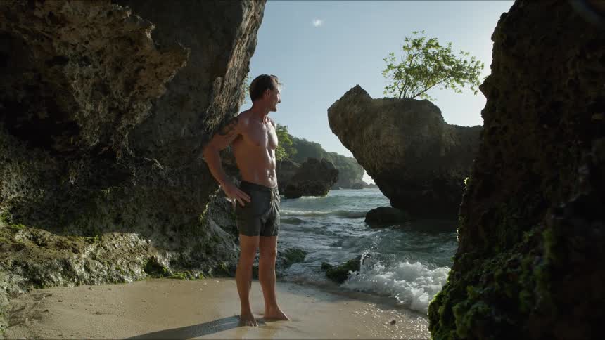 A man walking slowly on a shoreline beach stops to glance at his watch with concern