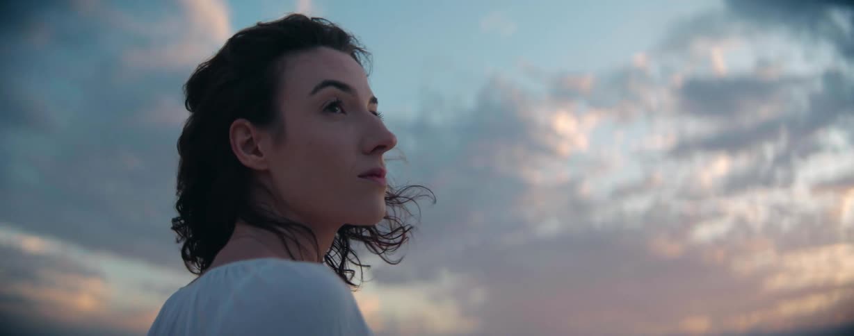 A woman with her hair flowing in the wind looks up at the clouds at sunset