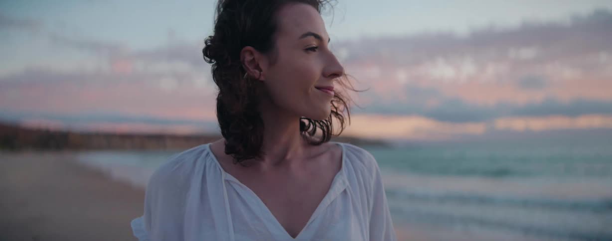 Woman with wavy hair and a white shirt walks along the sandy beach watching the sea at sunset