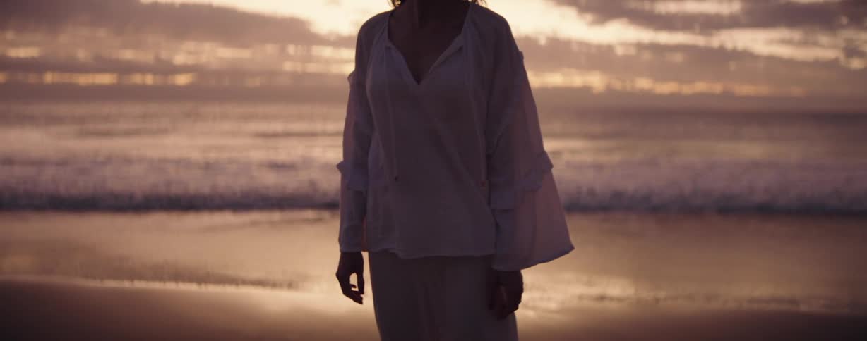 Woman in a white shirt and pants stands on the beach near the sea at sunset