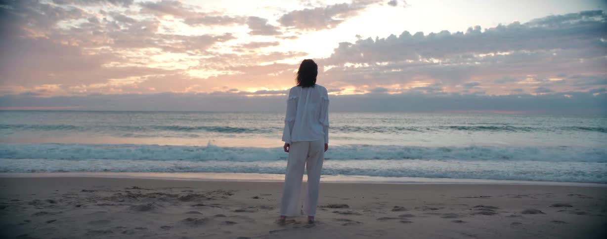 Woman wearing a white shirt and pants stands on the beach near the ocean at sunset