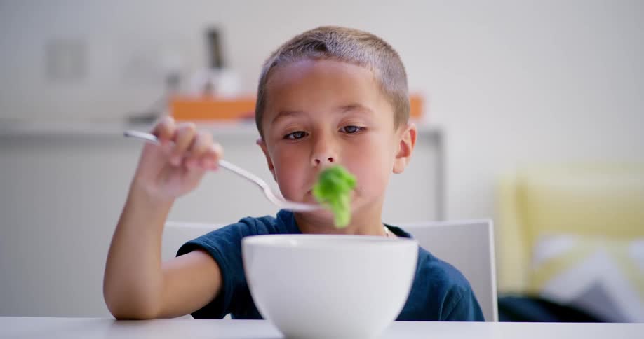 Disgusted young boy picking up a piece of broccoli from white bowl
