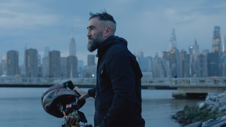 A man takes off a helmet and puts it on top of the motorcycle and then looks in front of him and behind him the water of a bay and behind him there is a city