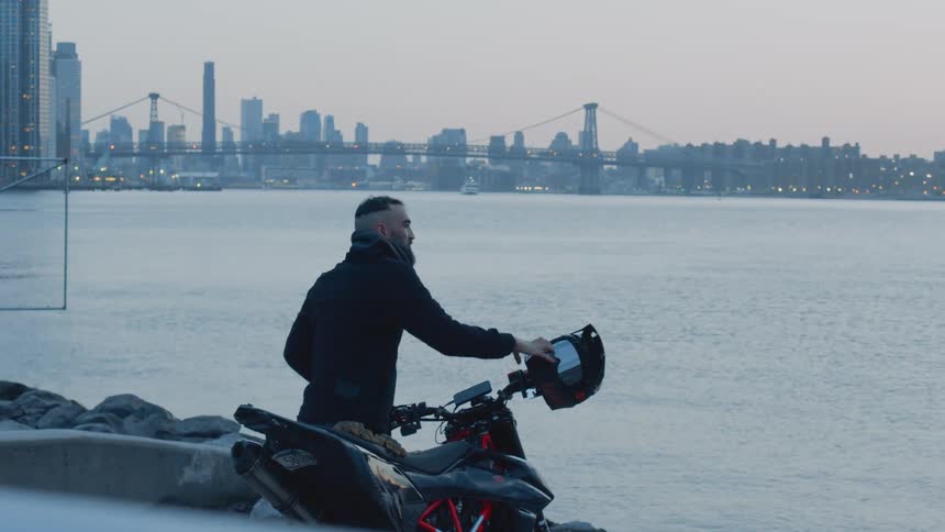 Man places helmet on motorcycle parked across water from downtown on a hazy day