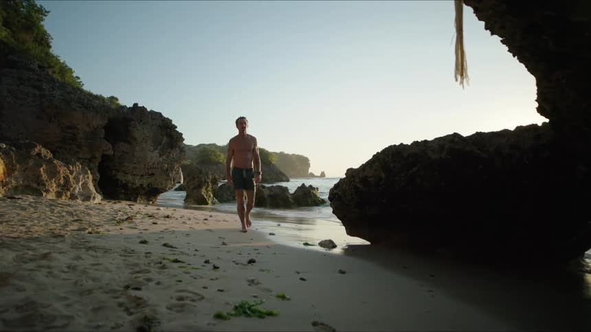 Man without a shirt and just shorts walking next to stones and rocks on a paradisiacal beach