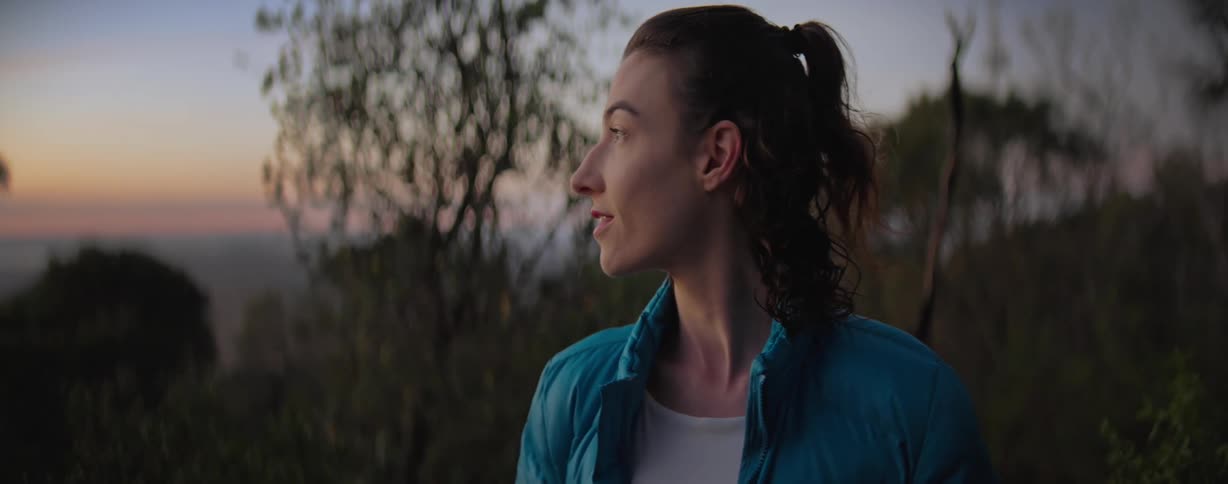 A female wearing a blue jacket is looking out from ontop of a mountain at sunrise