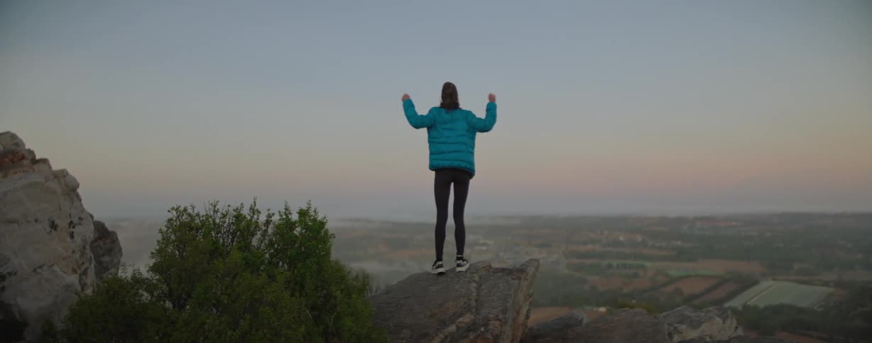 A woman in a blue jacket running to the edge of a cliff and raises her arms up