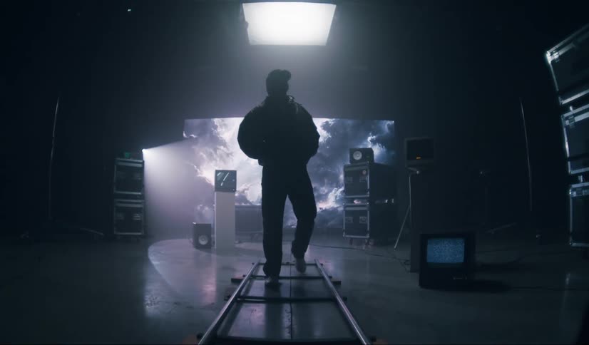 A male walking across a flat floor ladder in a dark video room that has monitors and sound equipment all over