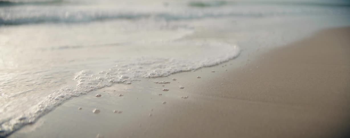 A beach with sand and small ripples of waves with some sea foam bubbles