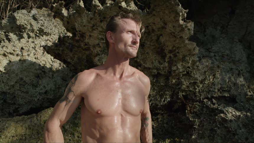 A muscled man with no shirt and arm tattoos is looking at his watch near craggy rocks