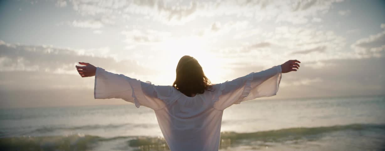 Woman wearing a white shirt stands on the beach with arms outstretched looking out at the ocean at sunset