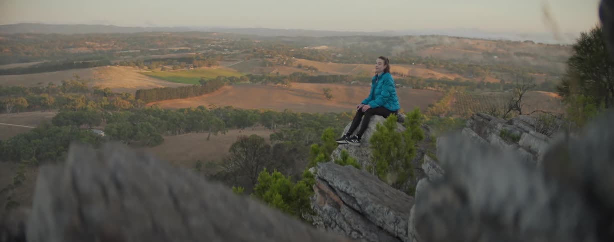A woman in a blue jacket sits on a rocky outcrop gazing out over a vast scenic landscape of rolling hills and fields at sunset