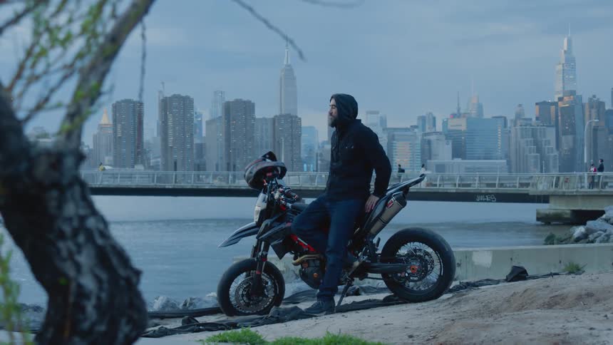 A person sitting on a motorcycle parked by a riverbank next to a city