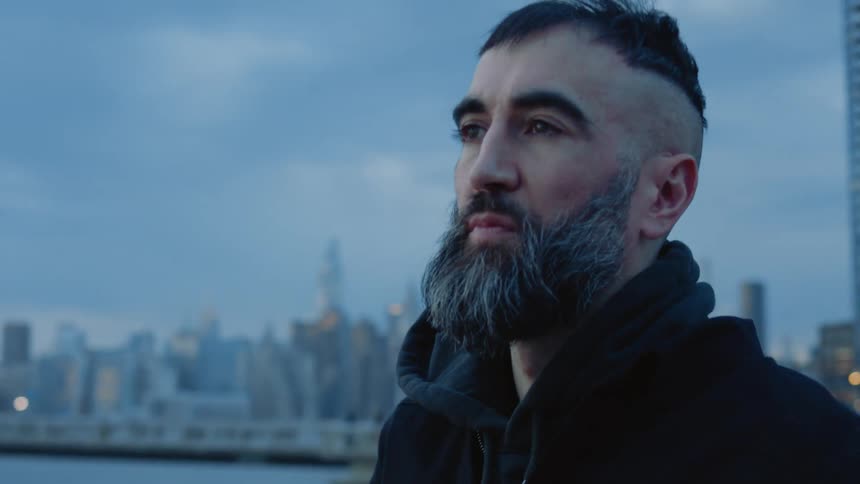 A bearded man takes a helmet off his head and looks ahead