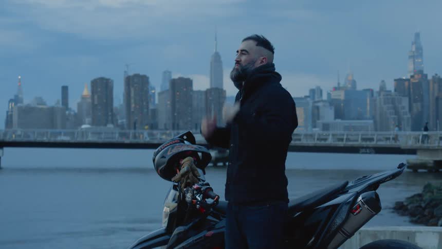 A man takes off his helmet and puts it on top of a motorcycle that is behind him and then puts his hands in his pocket and looks straight ahead