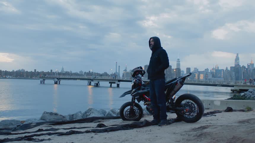 Man standing next to his motorbike and taking off his helmet and enjoying the seaside view with a metropolis in the background