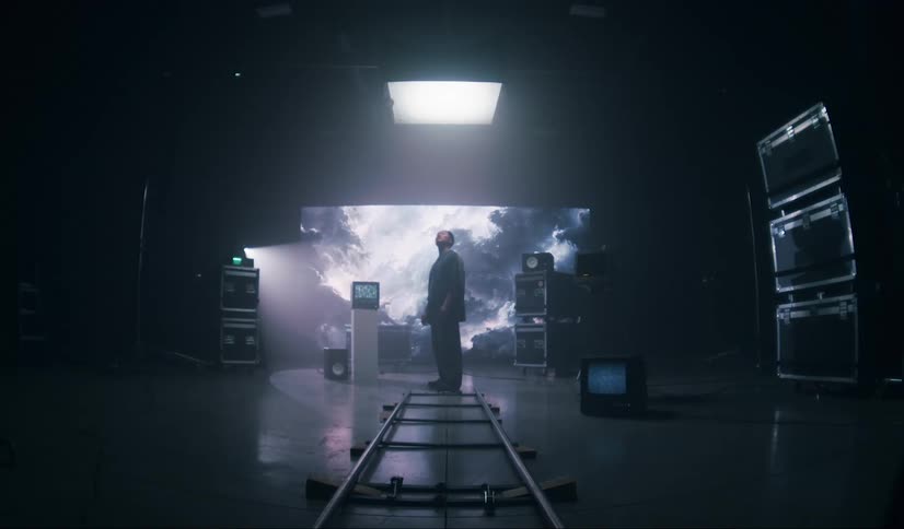 A person stands in a dark studio with a large light in the ceiling and a large screen behind