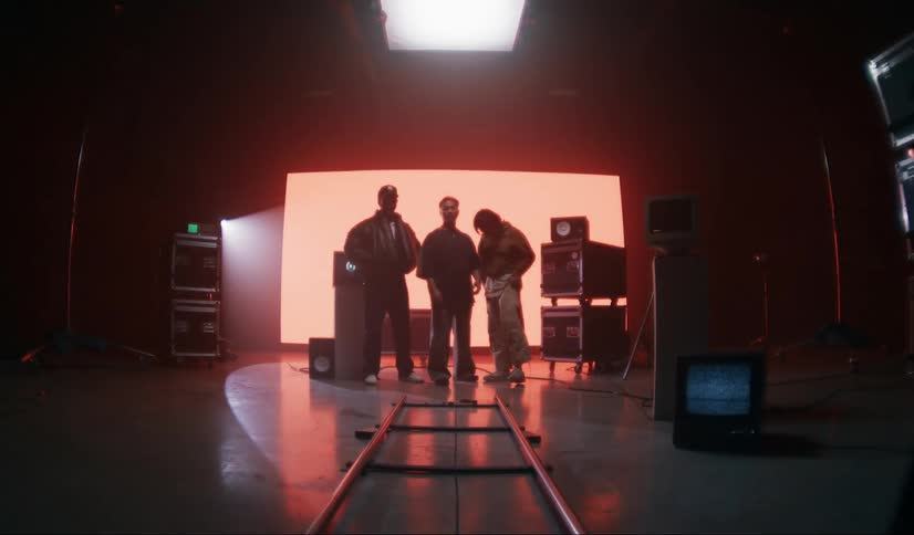 Three people stand in front of a large screen on a film set surrounded by camera equipment and lighting