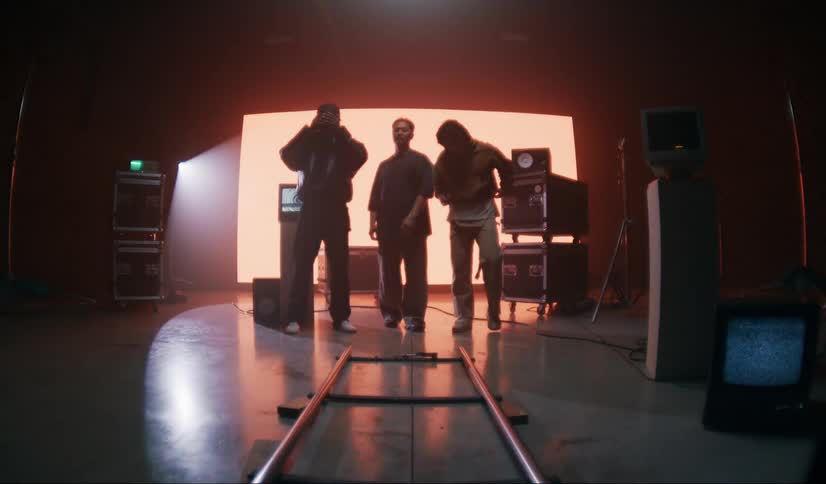 Three male models in a studio with old televisions and machines and a screen with red light in the background