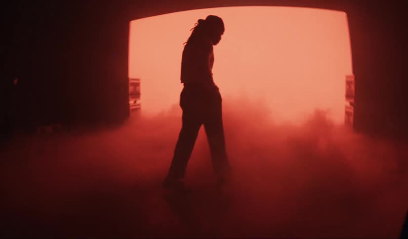A male with a hat and long hair is in a smoky room with red lighting and a large screen