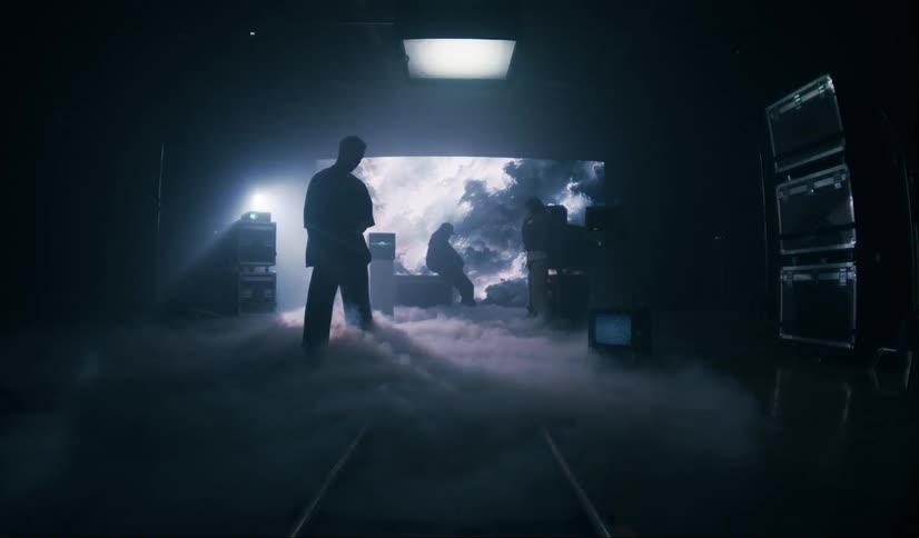 Men in a dark and hazy environment lit by a light suspended from the ceiling with a large screen in the background showing a hazy scene