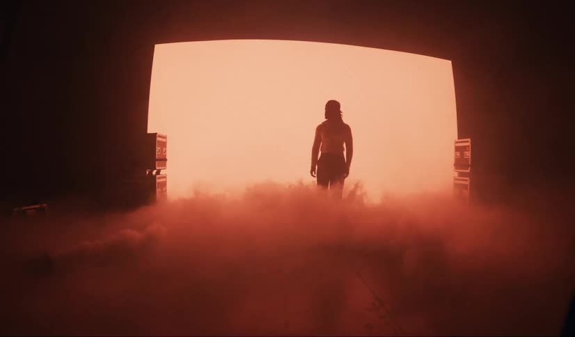A person walking in a place with the ground covered in smoke and red lights flashing in a black environment