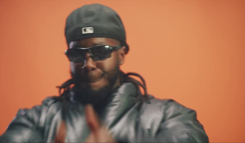 A man in a black coat wearing sunglasses and a cap is making movements with his hands and moving on an orange background