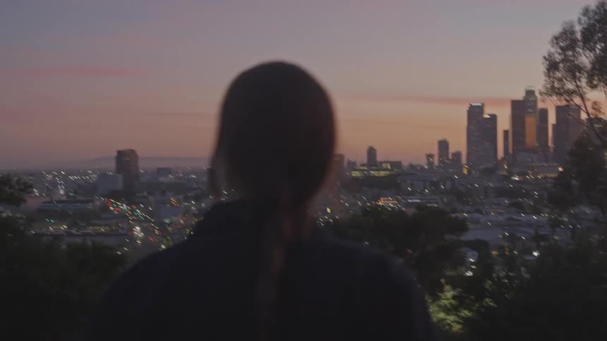 A female with a long braid standing next to a motorcycle is looking at a city horizon at dusk