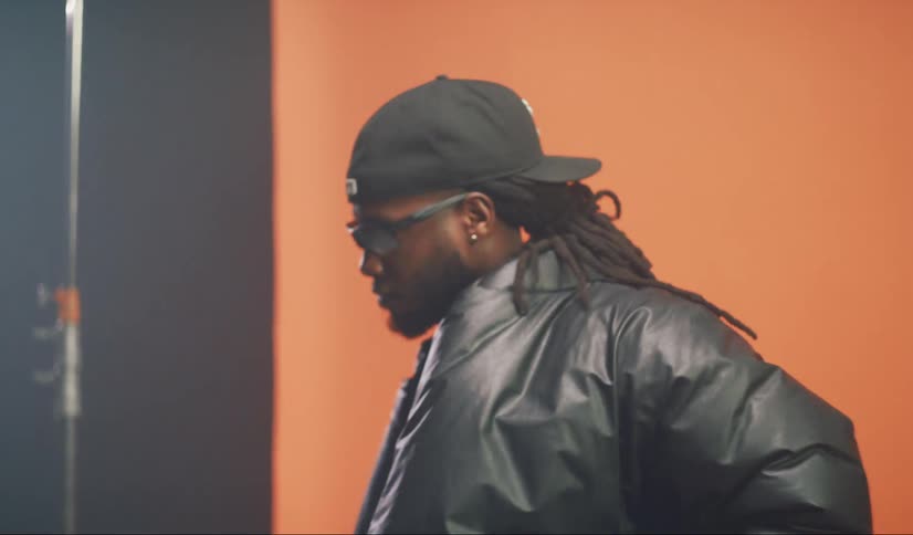 Man wearing a coat and a baseball cap bobs his head while moving back and forth near a red backdrop