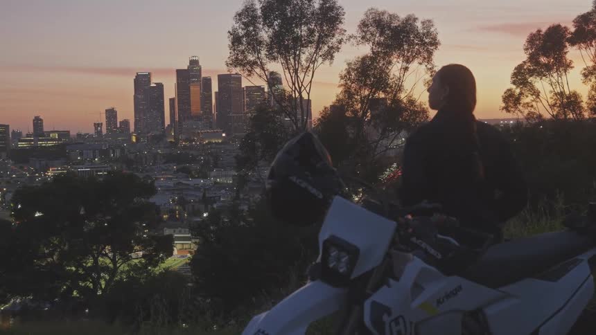 Woman leaning on a motorcycle against a sunset seen from a hill and metropolis in the background